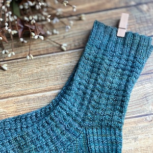 May include: A close-up of a teal blue knitted sock with a textured pattern. The sock is hanging from a wooden clothespin on a wooden surface.