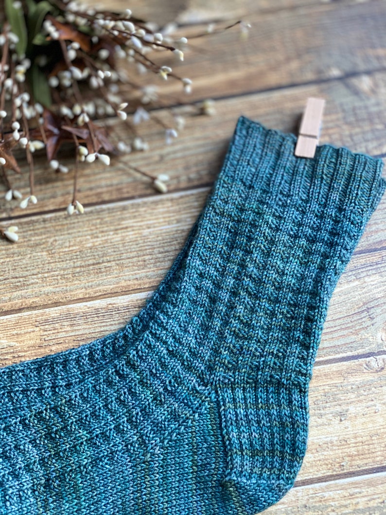 May include: A close-up of a teal blue knitted sock with a textured pattern. The sock is hanging from a wooden clothespin on a wooden surface.
