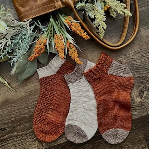 May include: Three pairs of knitted socks in a rust orange and beige colour scheme. The socks are made with a textured stitch and have a contrasting colour at the heel and toe.