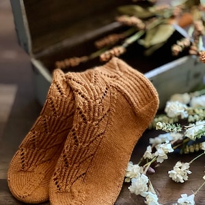 May include: A pair of brown knitted socks with a lacy pattern. The socks are lying on a wooden surface with white flowers.