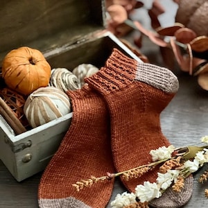 May include: A pair of knitted socks in a rust orange color with a brown cuff. The socks are lying on a wooden surface with dried flowers.