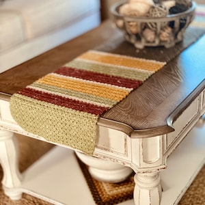 May include: A white coffee table with a distressed finish and turned legs. A crocheted table runner with stripes of green, yellow, brown, and red is on the table. A bowl of decorative balls is on the table.