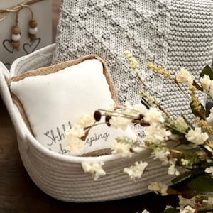 Puede incluir: Una cesta tejida blanca llena de una manta de punto gris, una almohada blanca con el texto "Shh... I'm sleeping", y flores blancas secas.
