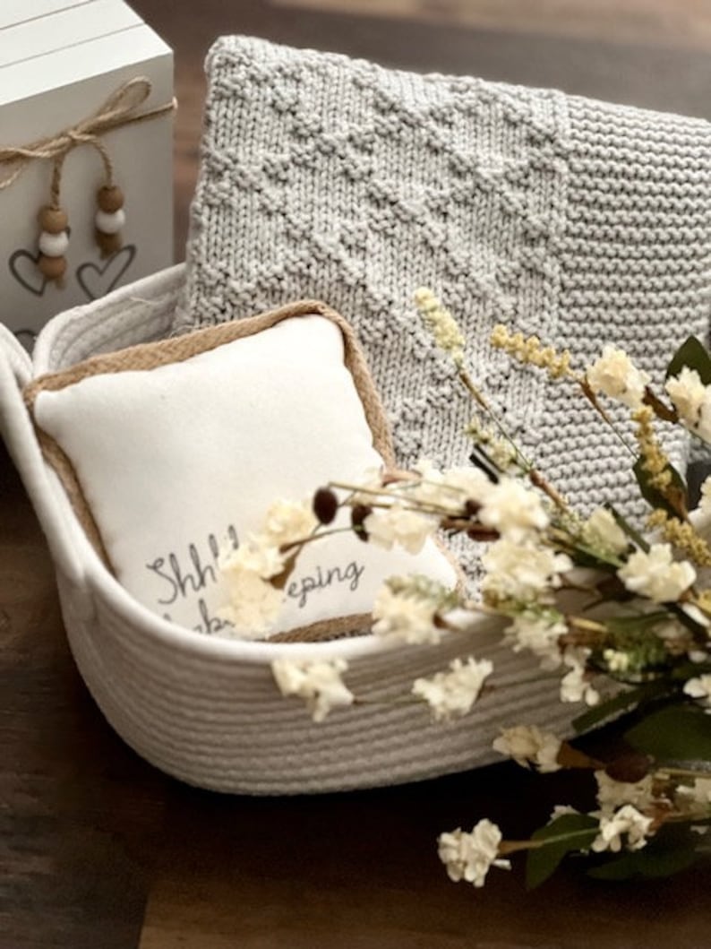 Puede incluir: Una cesta tejida blanca llena de una manta de punto gris, una almohada blanca con el texto "Shh... I'm sleeping", y flores blancas secas.