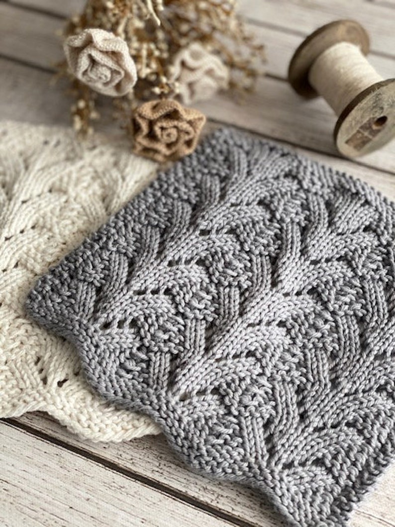 May include: Two knitted dishcloths, one white and one gray, with a delicate lace pattern. The gray dishcloth has a more intricate design.