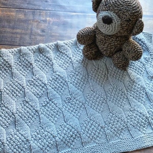May include: A light gray knitted baby blanket with a diamond pattern. A brown knitted teddy bear is sitting on top of the blanket.