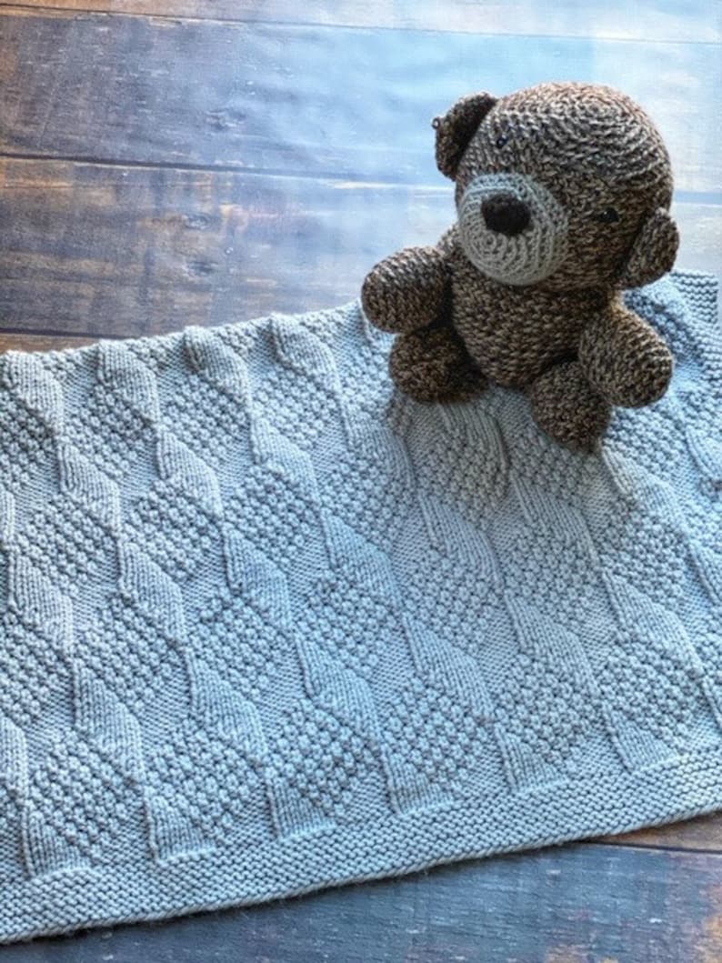 May include: A light gray knitted baby blanket with a diamond pattern. A brown knitted teddy bear is sitting on top of the blanket.