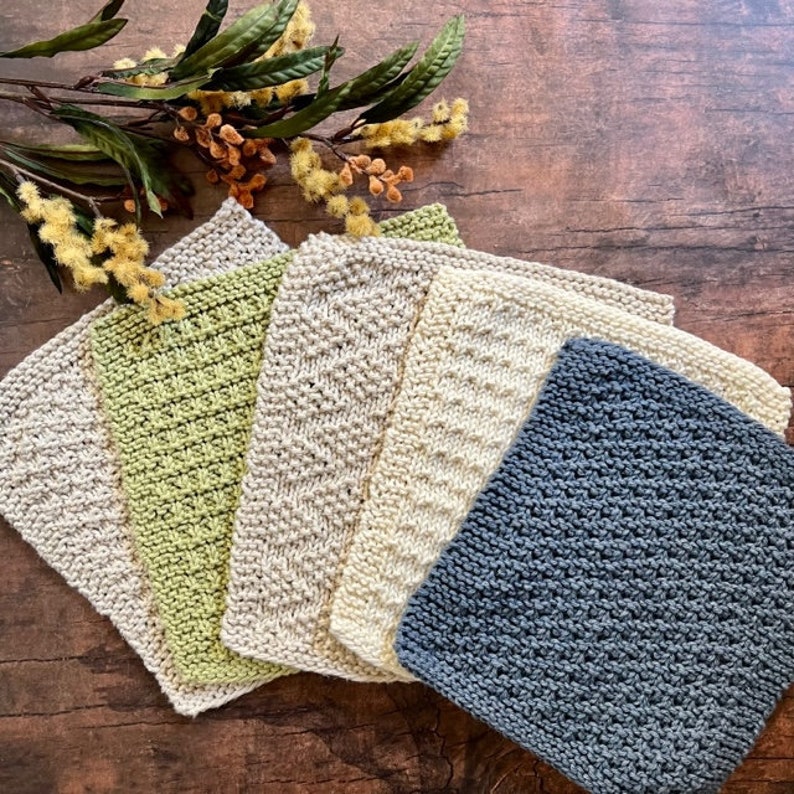 Pu&ograve; includere: Cinque strofinacci a maglia in vari colori e motivi. Gli strofinacci sono disposti a ventaglio su una superficie di legno. I colori sono verde, beige, crema e grigio.