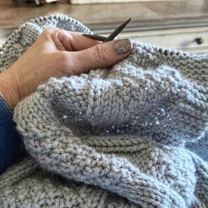 May include: A close-up of a person knitting a gray knitted blanket with gray knitting needles.