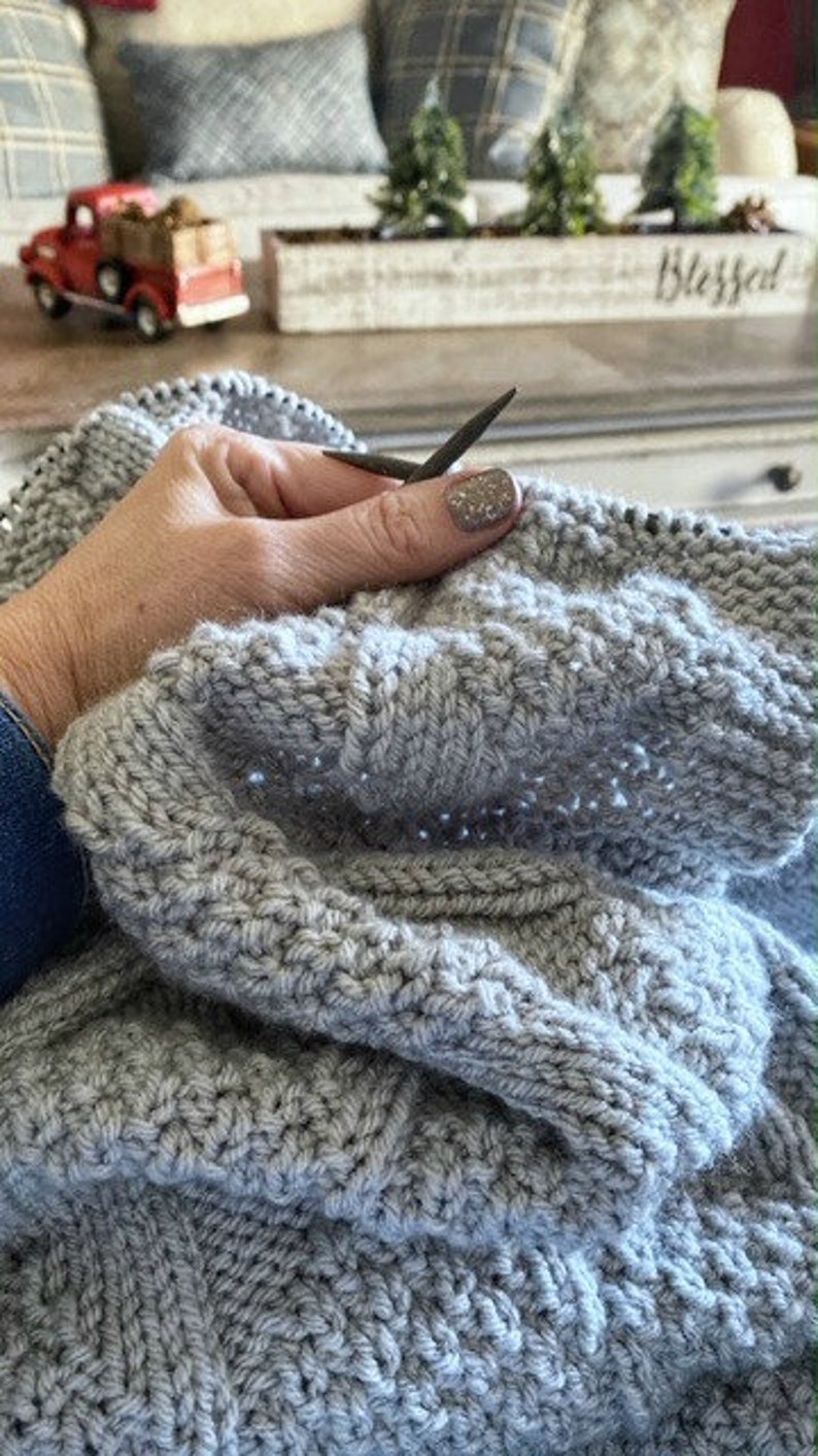 May include: A close-up of a person knitting a gray knitted blanket with gray knitting needles.