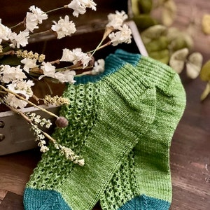 May include: A pair of green and teal knitted socks with a lacy pattern. The socks are lying on a wooden surface.
