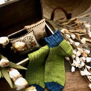 Könnte beinhalten: Ein Paar grün-blaue Stricksocken mit Rippendetails. Die Socken werden auf einer Holzoberfläche in der Nähe einer kleinen Holzkiste mit bestickten Kissen und Trockenblumen präsentiert. Die Socken haben blaue Fersen und Zehen.