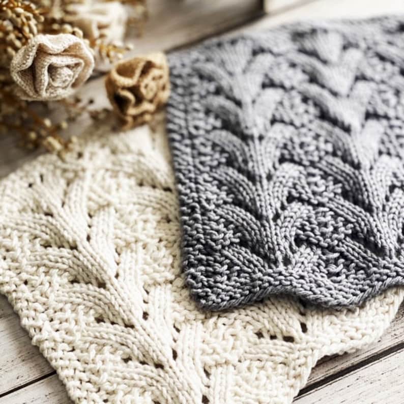 May include: Two knitted dishcloths, one white and one gray, with a cable knit pattern. The dishcloths are folded and placed on a wooden surface.