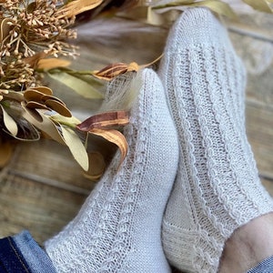 May include: A pair of white knitted socks with a cable knit pattern. The socks are worn on a person's feet and the person is wearing blue jeans.