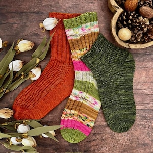 May include: Three pairs of knitted socks in shades of orange, green, and multicoloured stripes. The socks are arranged on a wooden surface with dried flowers and a wooden bowl filled with pine cones and nuts. The socks appear to be handmade.