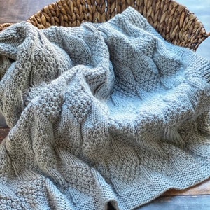 May include: A gray knitted baby blanket with a cable knit pattern. The blanket is folded and placed in a woven basket.