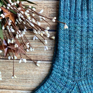 May include: A close-up of a teal blue knitted sock with a ribbed cuff. The sock is made with a textured yarn and has a subtle speckled pattern.