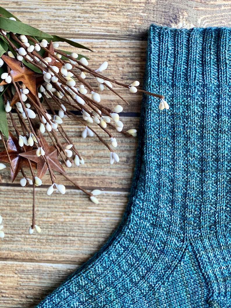 May include: A close-up of a teal blue knitted sock with a ribbed cuff. The sock is made with a textured yarn and has a subtle speckled pattern.
