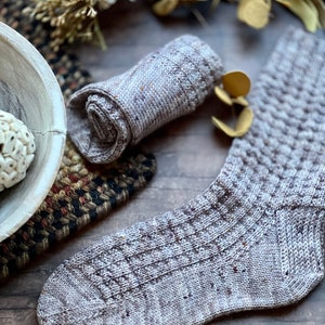 May include: A pair of knitted socks in a light brown and grey color with a textured pattern. The socks are rolled up and laid out on a wooden surface.