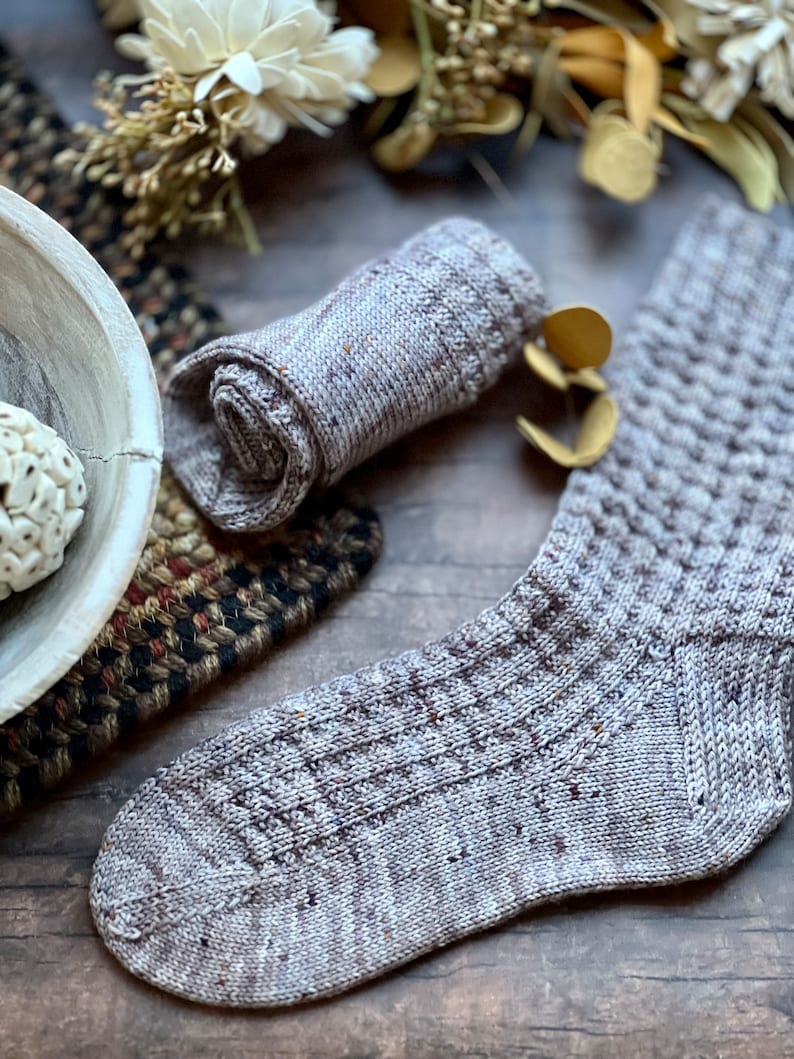 May include: A pair of knitted socks in a light brown and grey color with a textured pattern. The socks are rolled up and laid out on a wooden surface.