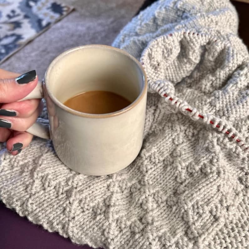 Puede incluir: Una taza de cer&aacute;mica blanca con asa, llena de caf&eacute;, descansando sobre una manta de punto gris con un hilo rojo que recorre el borde.