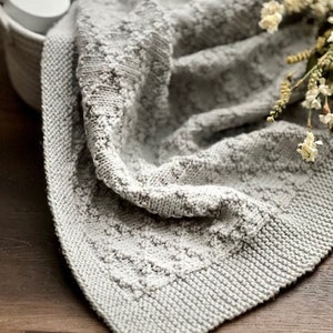 Puede incluir: Una suave manta de beb&eacute; de punto gris con un patr&oacute;n texturizado. La manta est&aacute; doblada y drapeada sobre una superficie de madera.