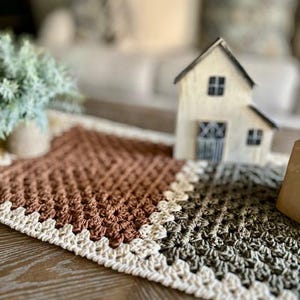 May include: A crocheted table runner with a brown, white, and green geometric pattern. The runner has a white crocheted border.