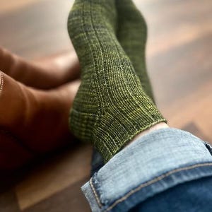 Könnte beinhalten: Ein Paar grüne Strickstrümpfe mit Rippenstruktur, getragen mit blauen Jeans. Die Socken sind eine Mischung aus grünem und schwarzem Garn, und die Jeans haben eine ausgewaschene Optik. Die Person trägt braune Lederstiefel.