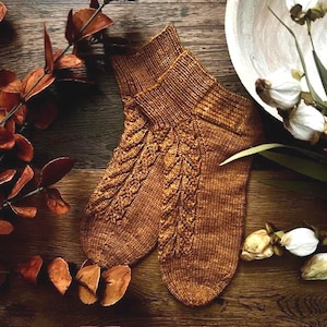 May include: A pair of knitted socks in a warm brown hue, featuring a textured leaf pattern along the front. The socks have ribbed cuffs and are displayed on a wooden surface with dried leaves and a white bowl with flowers.