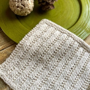 May include: A close-up of a knitted dishcloth in a light beige color. The dishcloth is made with a simple stitch pattern and has a slightly textured surface.