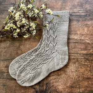 May include: A pair of grey knitted socks with a lacy pattern. The socks are lying on a wooden surface.