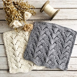May include: Two knitted dishcloths, one white and one gray, with a leaf pattern. The dishcloths are laid out on a white wooden surface.