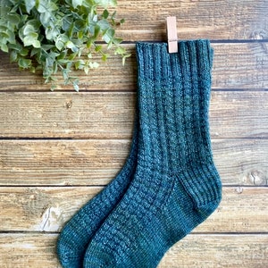 May include: A pair of teal blue knitted socks with a ribbed texture. The socks are hanging on a wooden surface with a clothespin.