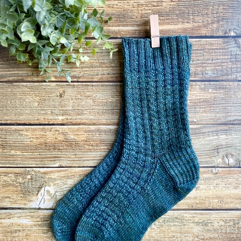 May include: A pair of teal blue knitted socks with a ribbed texture. The socks are hanging on a wooden surface with a clothespin.