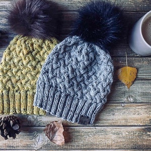 May include: Two knitted winter hats with faux fur pom poms. One hat is yellow and the other is gray. The hats are on a wooden surface with a cup of coffee and fall leaves.