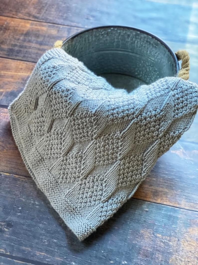 May include: A gray knitted blanket with a cable knit pattern. The blanket is folded and draped over a metal bucket.
