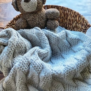 May include: A light blue knitted baby blanket with a brown crocheted teddy bear on top of it. The blanket is in a wicker basket.