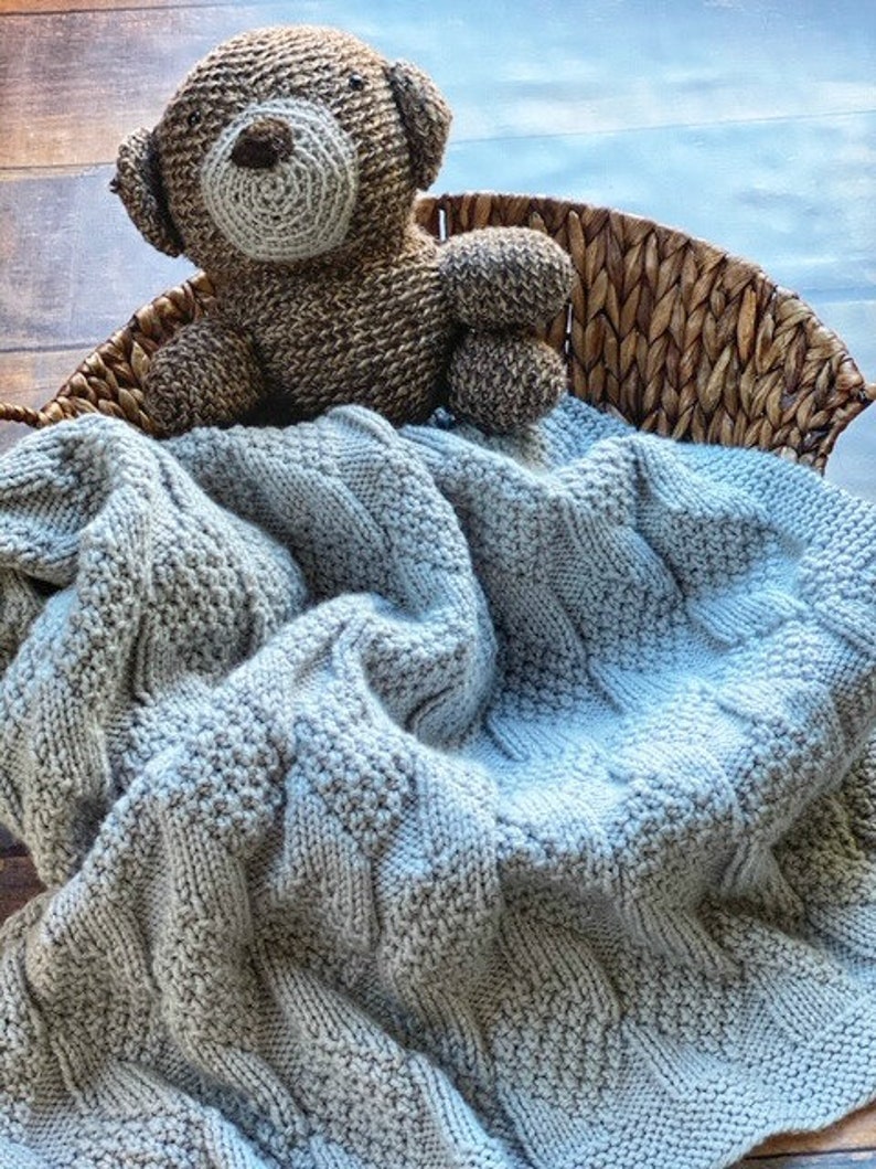 May include: A light blue knitted baby blanket with a brown crocheted teddy bear on top of it. The blanket is in a wicker basket.