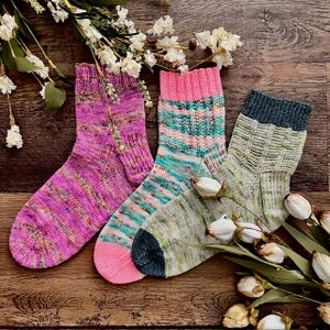 May include: Three pairs of colorful, hand-knitted socks. One pair is primarily pink with a speckled pattern, another has pink, teal, and white stripes, and the third is green with a textured design. The socks are arranged on a wooden surface.