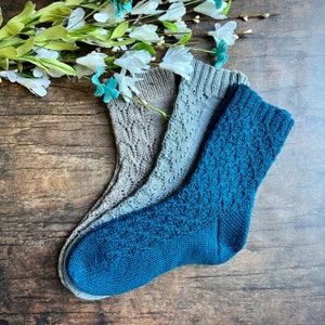 May include: Three pairs of knitted socks, one pair is teal blue, and two pairs are a light gray color. The socks are laid out on a wooden surface.
