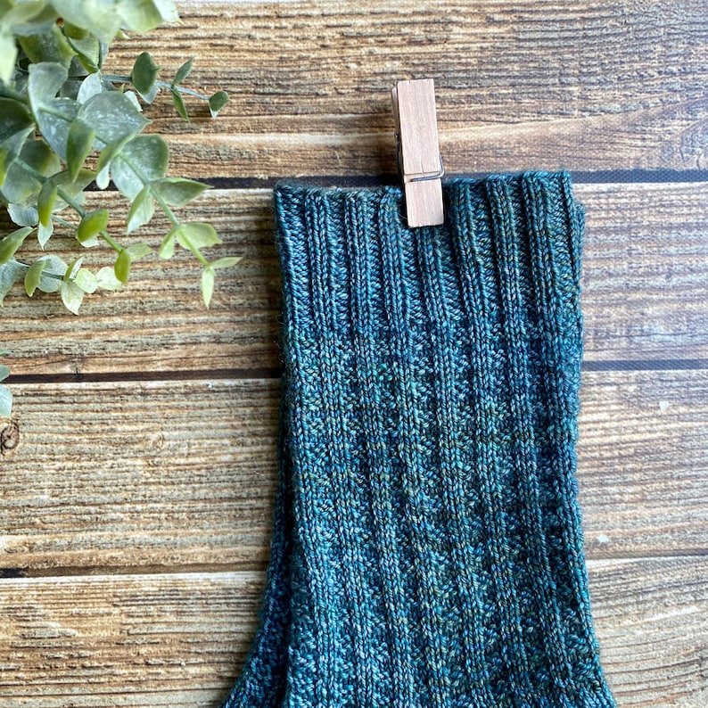 May include: A pair of teal blue knitted socks with a ribbed texture. The socks are hanging from a wooden clothespin.