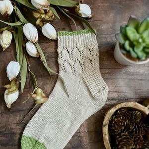 May include: A pair of beige knitted socks with a green trim. The socks have a lacy pattern and are photographed on a wooden surface.