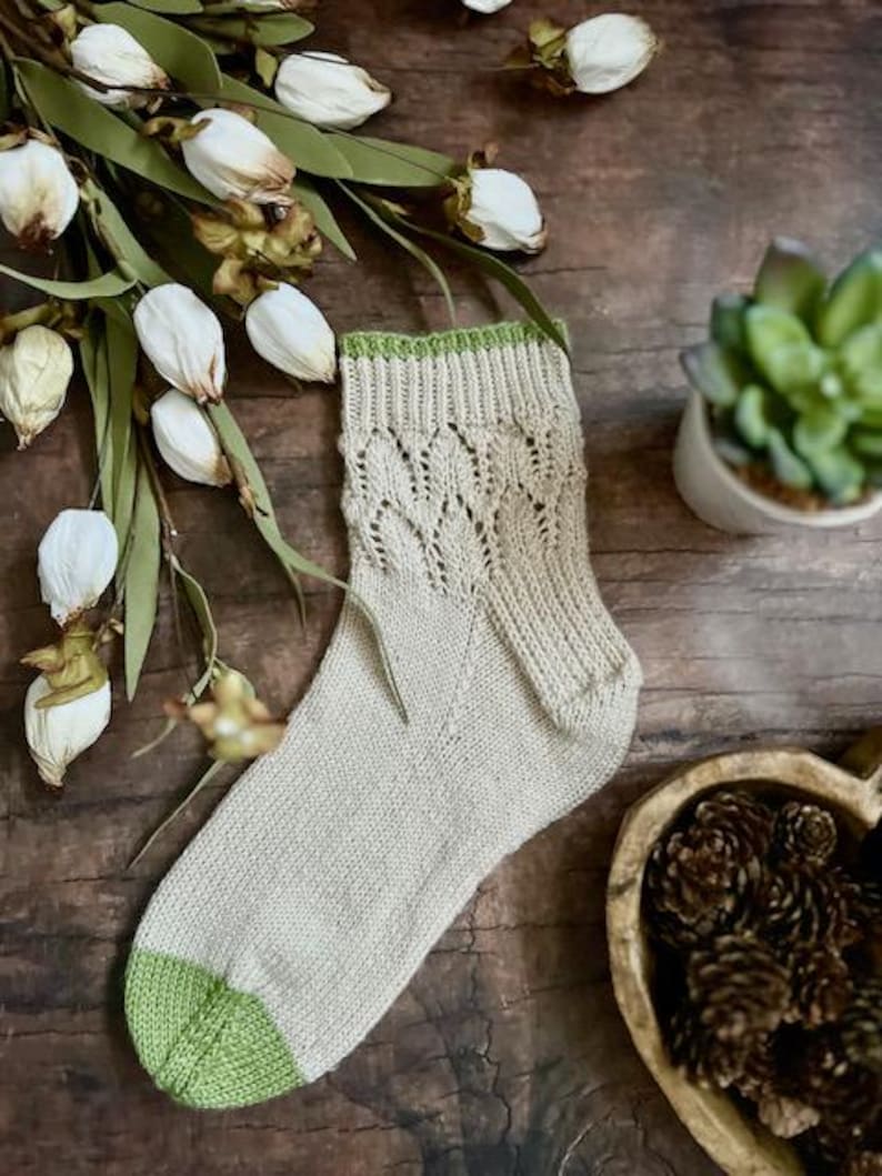 May include: A pair of beige knitted socks with a green trim. The socks have a lacy pattern and are photographed on a wooden surface.
