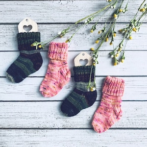 May include: Three pairs of hand-knitted socks in pink and green color combinations. The socks are hanging on wooden tags with heart cutouts.