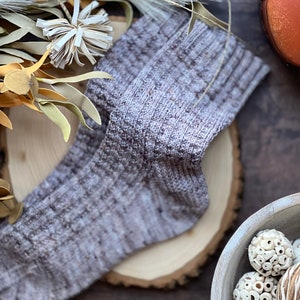 May include: A pair of gray and brown knitted socks with a textured pattern. The socks are lying on a wooden surface.