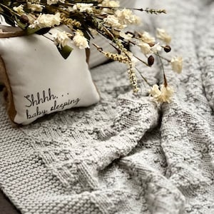 Puede incluir: Una manta de beb&eacute; de punto gris con una almohada blanca que dice "Shhhh... beb&eacute; durmiendo". La manta es texturizada y tiene un patr&oacute;n de punto de cable. Un ramo de flores blancas secas est&aacute; encima de la almohada.