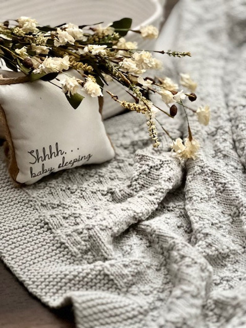 Puede incluir: Una manta de beb&eacute; de punto gris con una almohada blanca que dice "Shhhh... beb&eacute; durmiendo". La manta es texturizada y tiene un patr&oacute;n de punto de cable. Un ramo de flores blancas secas est&aacute; encima de la almohada.