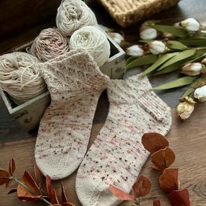 May include: Hand-knitted socks with a speckled pattern in cream, pink, and brown. The socks are displayed with yarn balls in a wooden box and autumn decorations, creating a cosy, handcrafted aesthetic.