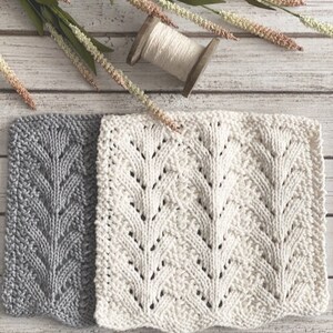May include: Two knitted dishcloths, one gray and one white, with a leaf pattern. The dishcloths are laid out on a white wooden surface.