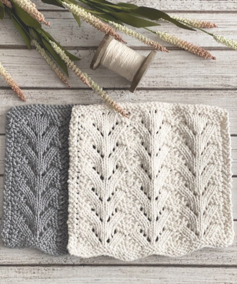 May include: Two knitted dishcloths, one gray and one white, with a leaf pattern. The dishcloths are laid out on a white wooden surface.